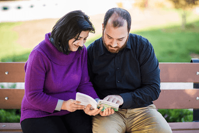 A man and woman sitting on a park bench reading a book together, both focused and smiling slightly.